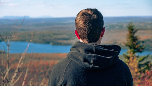 A man with a hooded jacket resting on a hill looking at  a beautiful river landscape on a sunny day