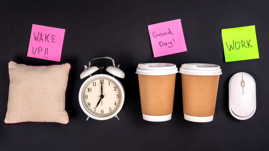 Paper cups, an alarm clock, a computer mouse and bright paper stickers, on a dark background, top view. The concept of morning at work, coffee in the office, coffee addiction.