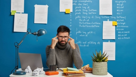 Unhappy male worker in spectacles sits at desktop during hard workday, keeps fingers on temples, suffers from headache, tries to concentrate on object, tired of overworking time at laptop computer